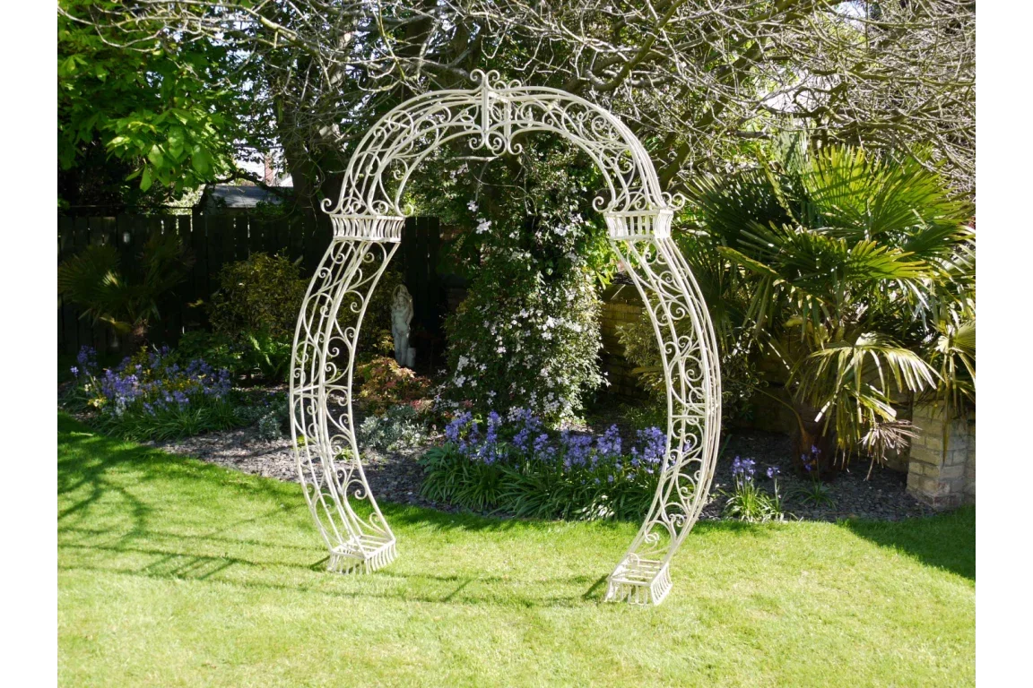 Metal garden arch with ornate scrollwork on grass lawn surrounded by plants and trees