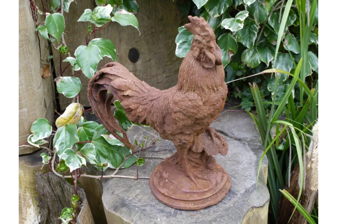 Rustic metal rooster garden ornament on wooden log surrounded by green plants and leaves