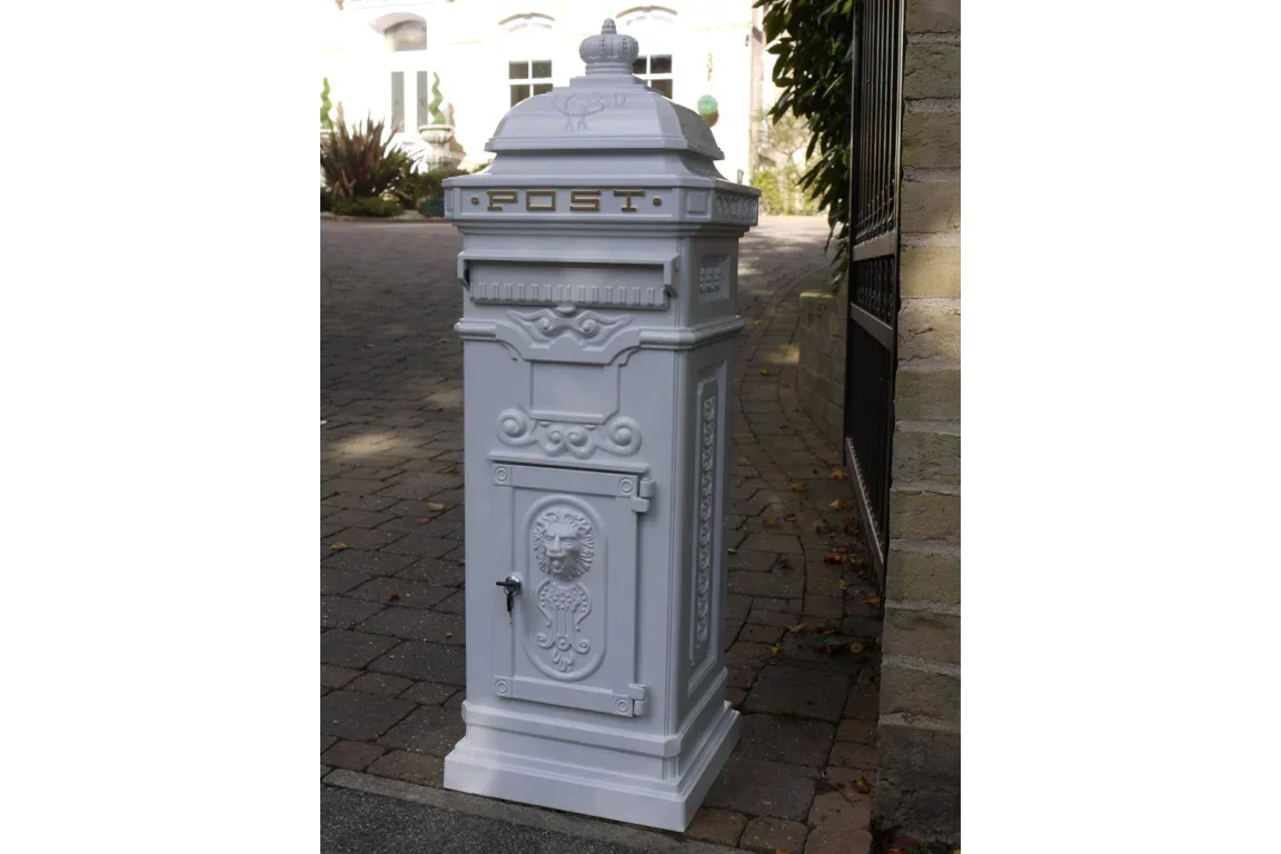 White cast iron post box with ornate detailing on stone driveway by iron gate