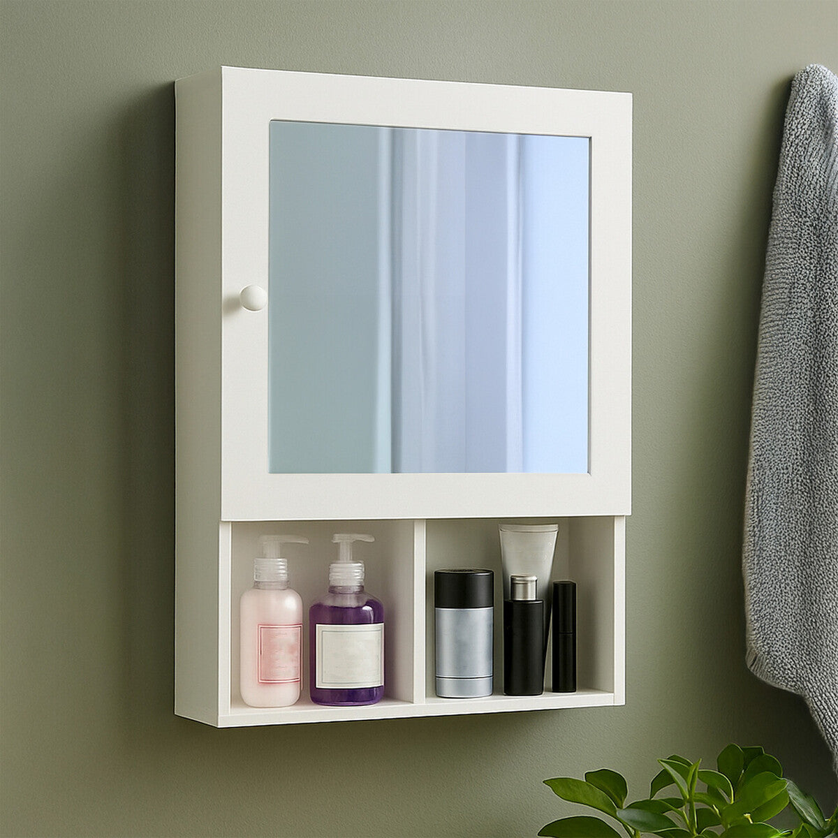 White wood cabinet with a mirrored front, featuring two mirrored doors and an open shelf below. The cabinet is mounted on a green wall and displays various bottles and containers on the shelf. A gray towel hangs nearby