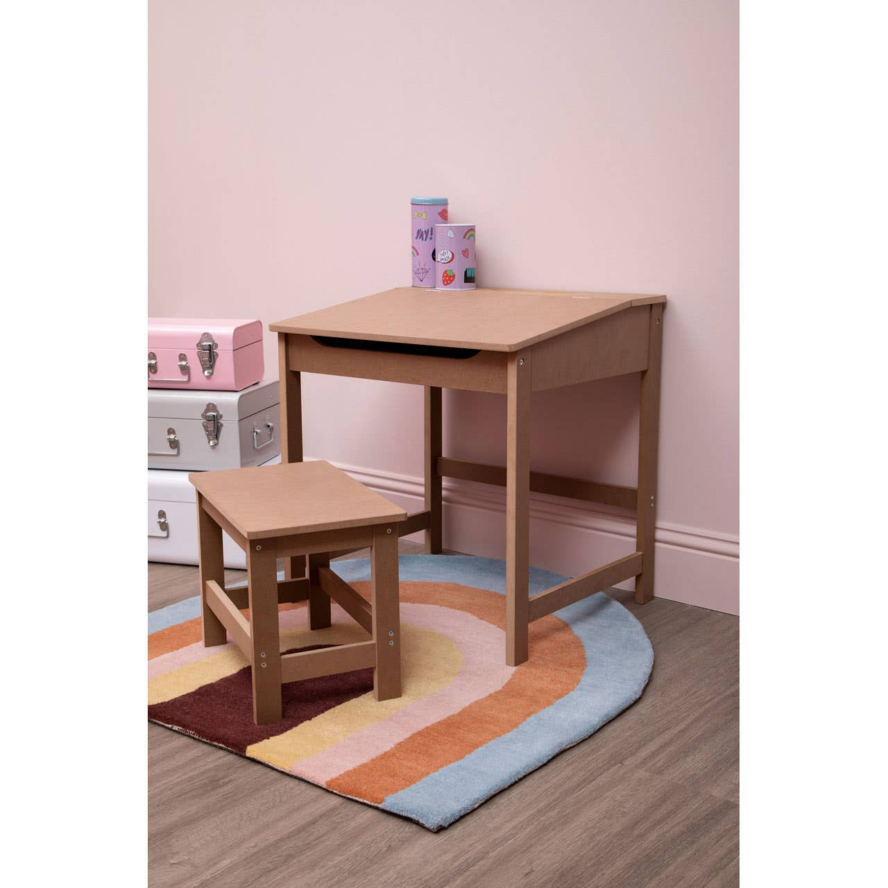 Natural wood desk and matching stool set for children, positioned on a colorful, circular rug. The desk features a simple, rectangular design with clean lines, while the stool is smaller and complements the desk's height. The background includes a soft pink wall, enhancing the playful atmosphere