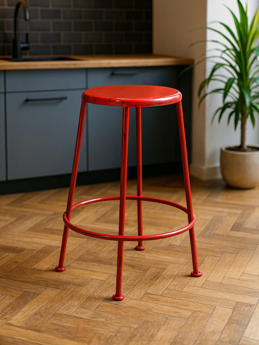 A bright red metal stool with a round seat and slender legs, positioned on a wooden floor in a modern kitchen setting with gray cabinetry and a potted plant in the background