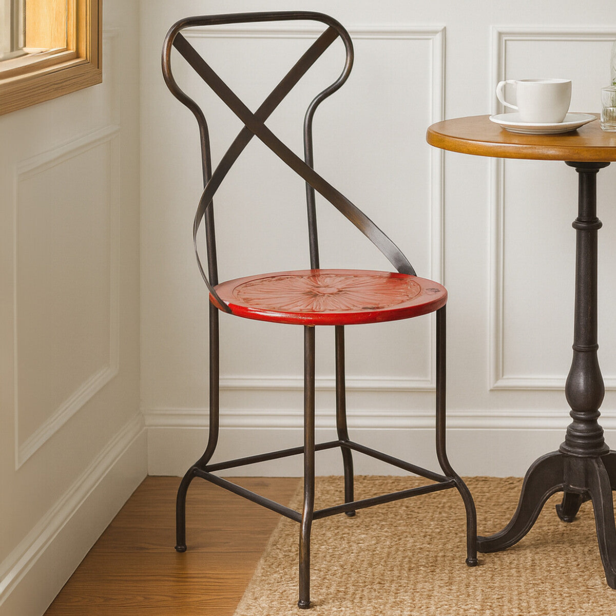A red metal seat chair with a cross-back design, positioned next to a small wooden side table. The chair features a round seat and slender black legs, set against a light-colored wall and wooden floor