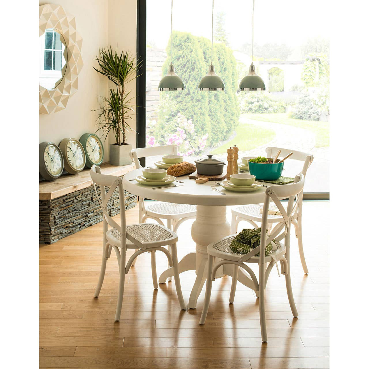 Round dining table with a white wash finish surrounded by four matching chairs, set in a bright room with large windows. The table is adorned with a decorative centerpiece, bowls, and green plants. A stylish mirror and decorative cushions are visible in the background, along with wooden flooring and pendant lights hanging above