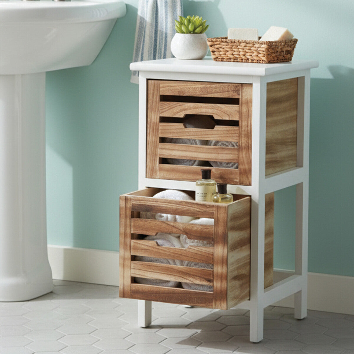 Two-drawer chest featuring a white frame and natural wood accents, with slatted design on the drawers. The top drawer is slightly smaller and has a decorative basket on top, while the bottom drawer is larger and open, displaying toiletries. The chest stands on four legs against a light blue wall, with a sink visible in the background