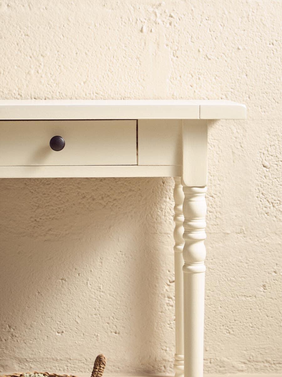 Cream wooden console table with black handle and turned leg detail against textured wall