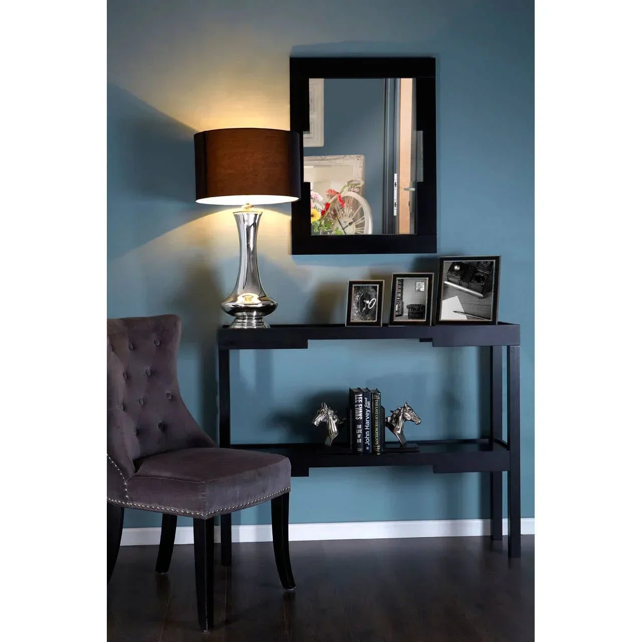 Black console table with silver lamp, framed photos, books, and grey upholstered chair in hallway
