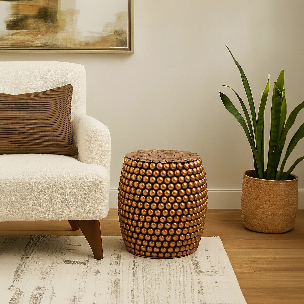 A copper finish barrel stool with a textured surface sits next to a cream-colored sofa adorned with a brown pillow. A potted plant with long green leaves is positioned nearby, and the scene is set on a light-colored area rug with a subtle pattern