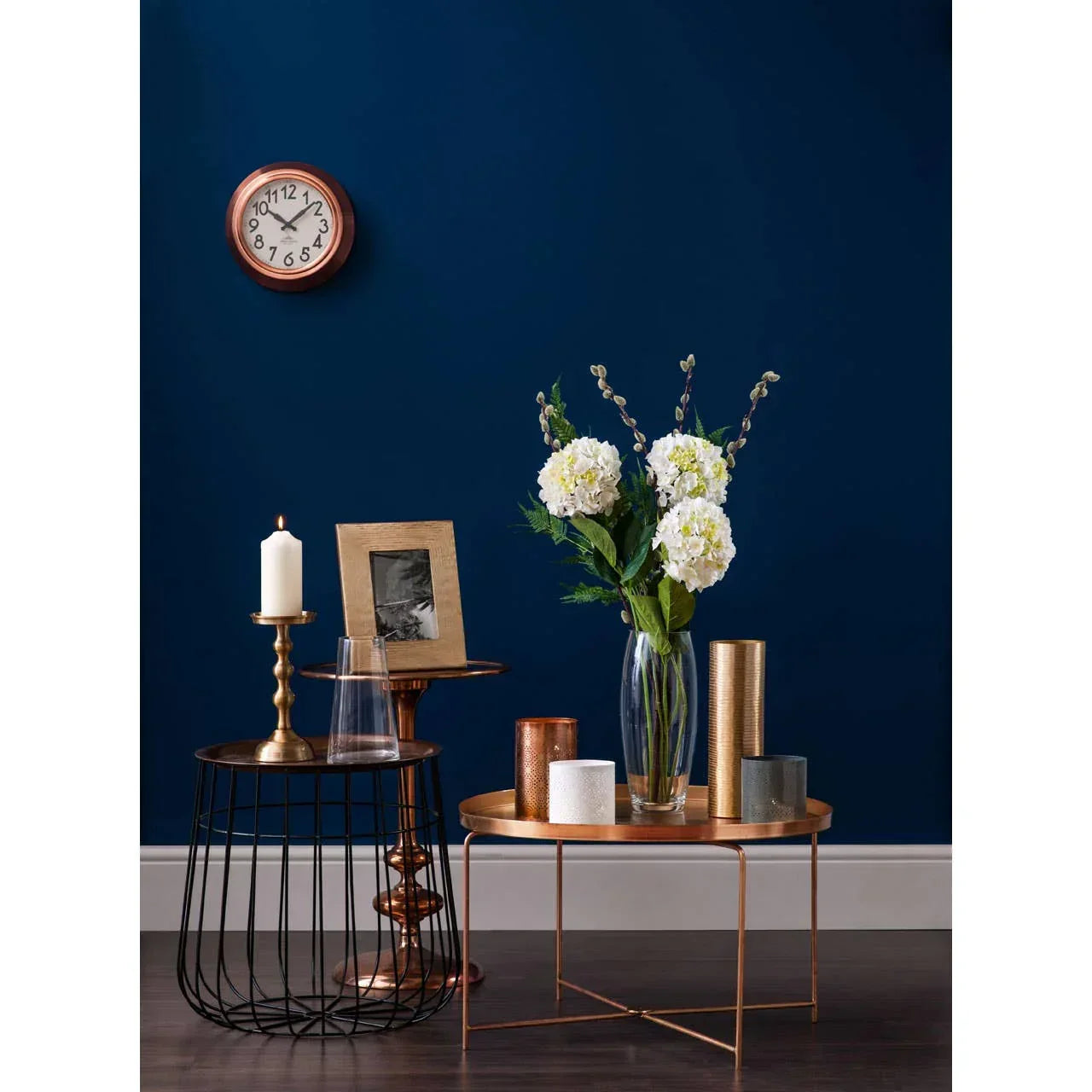 Copper metal side tables with gold candle holders, vase of white flowers, and wall clock