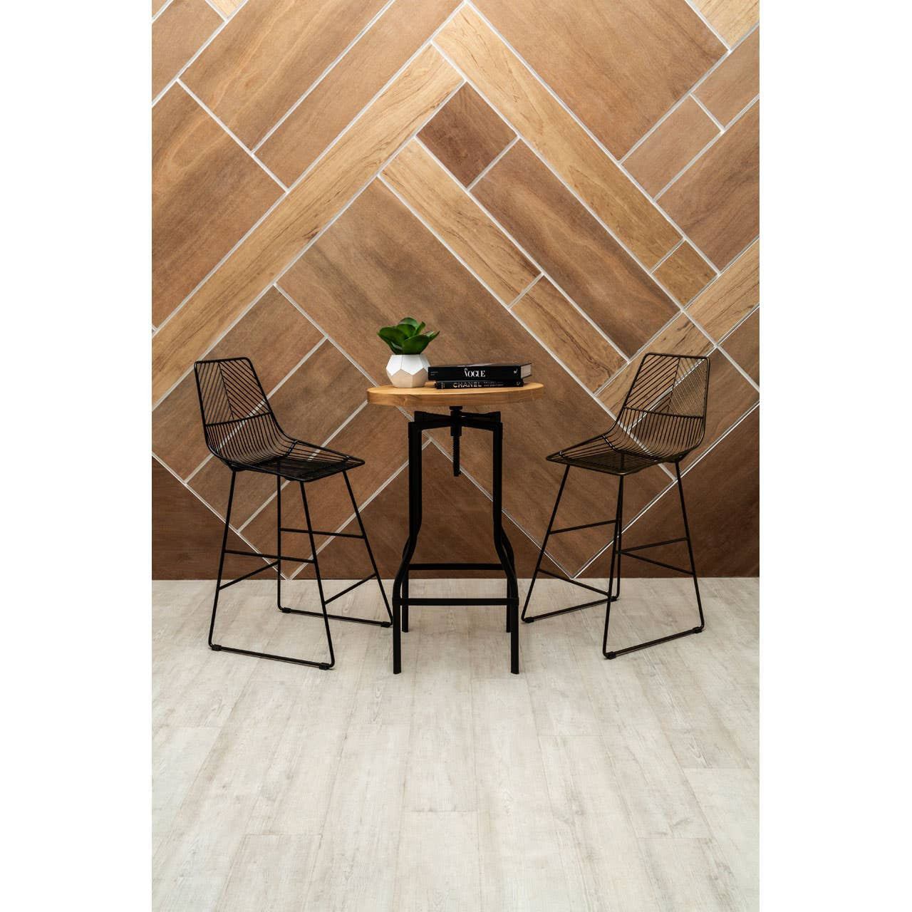 A bar table with a smooth ash veneer top and a sleek black metal base, accompanied by two wire-frame bar stools, set against a geometric wooden wall design. A small potted plant and a decorative item are placed on the table, with light-colored flooring visible beneath