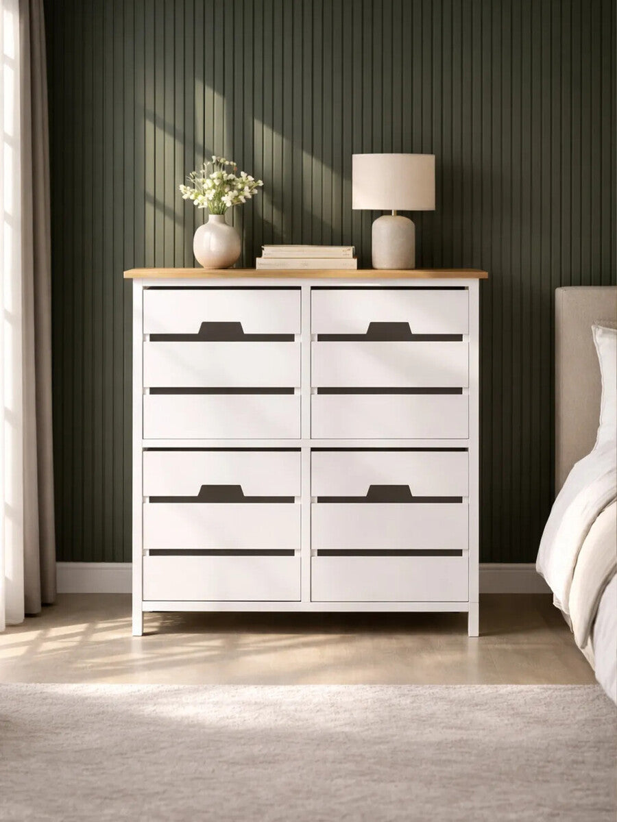 A white four-drawer chest with a natural wood top, featuring horizontal slats on the drawer fronts and black handles. The chest is positioned against a dark green wall, with a bedside lamp and a small vase of flowers on top, and a light-colored rug visible on the floor