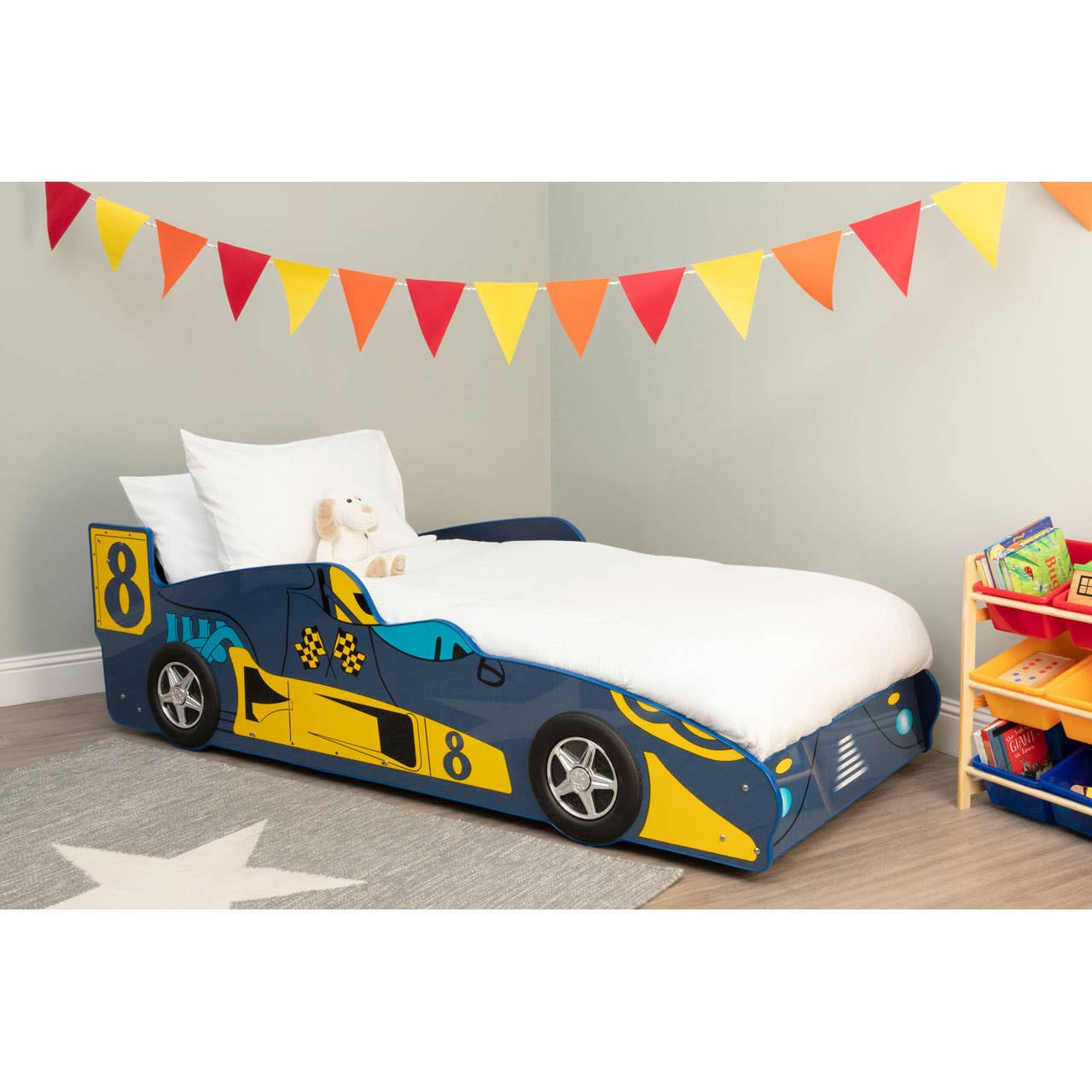 A colorful kids racing car bed designed to resemble a race car, featuring a blue and yellow color scheme with racing decals. The bed is positioned against a light gray wall, surrounded by festive triangular bunting in red, yellow, and orange. A white mattress with a pillow is placed on the bed, and a toy storage unit filled with colorful toys is visible in the background. A star-shaped rug is