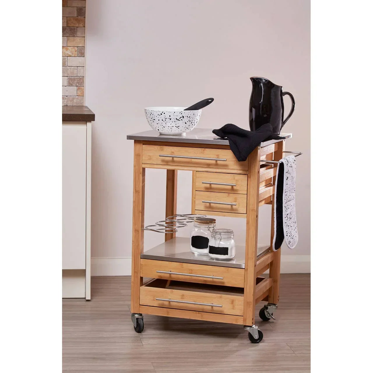 Wooden kitchen trolley with stainless steel top, drawers, shelves and black jug on top