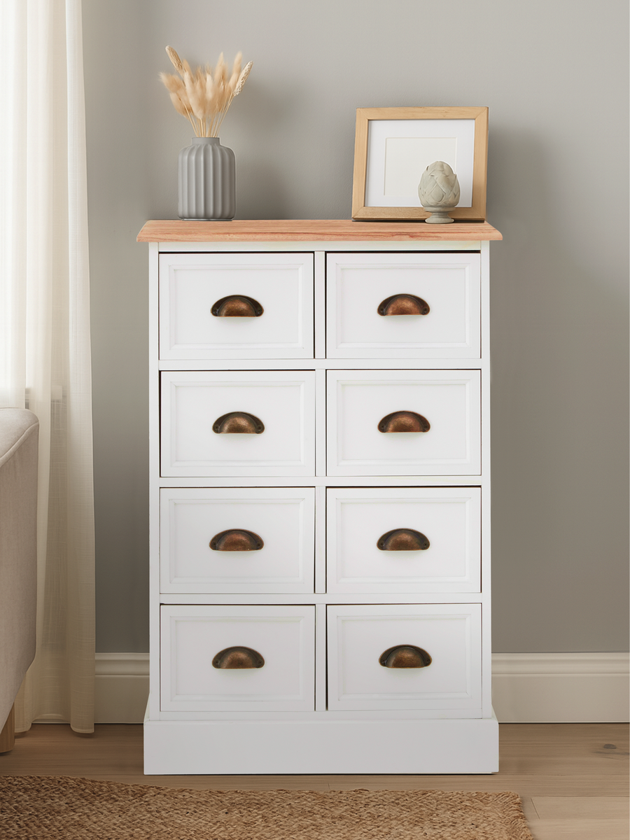 A white chest of drawers with eight drawers, featuring a natural wood top and simple cut-out handles, placed against a light gray wall. A small decorative frame and a vase with dried flowers are positioned on the top surface