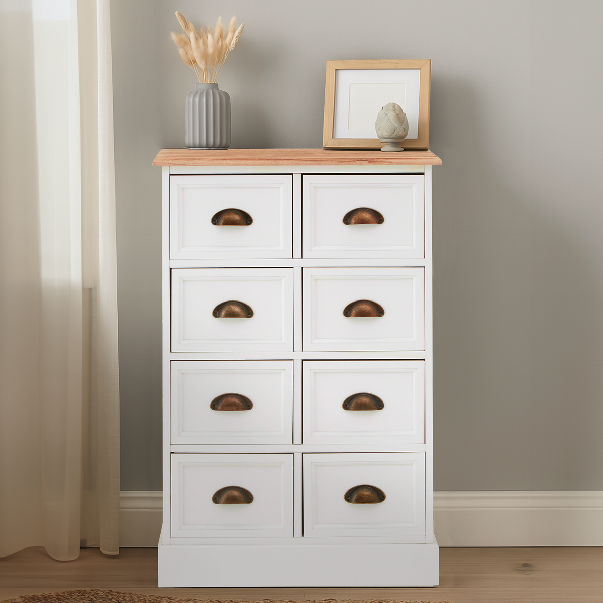 A white chest of drawers with eight drawers, featuring a light wood top and bronze handles, placed against a light gray wall. A small picture frame and a decorative vase with dried flowers are on top of the chest