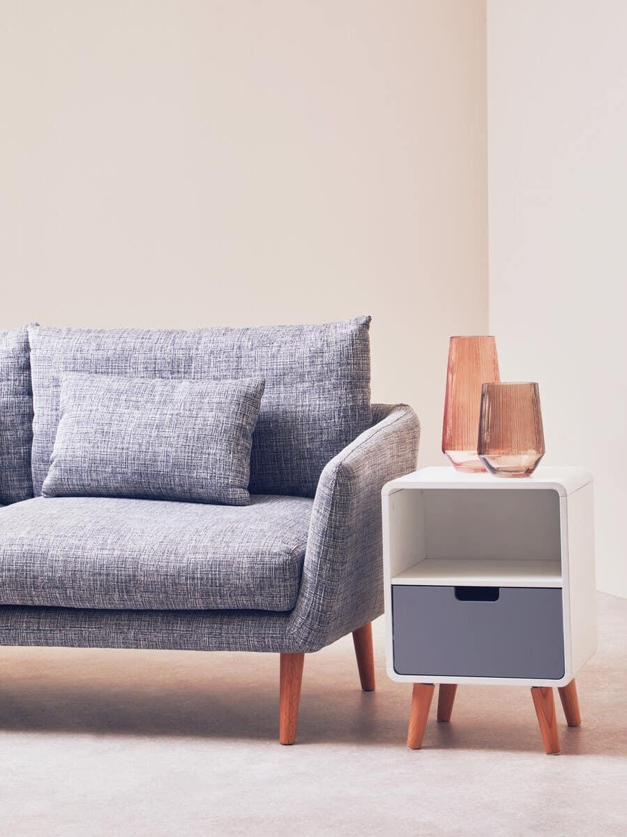 Grey fabric sofa with wooden legs next to modern white bedside table and glass vases