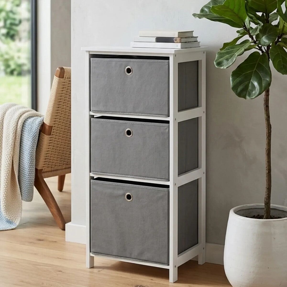 Three grey fabric drawers in a white cabinet with a modern design, placed against a light-colored wall. A potted plant is visible beside the cabinet, and a cozy throw blanket is draped over a nearby chair. The floor is wooden, adding warmth to the scene