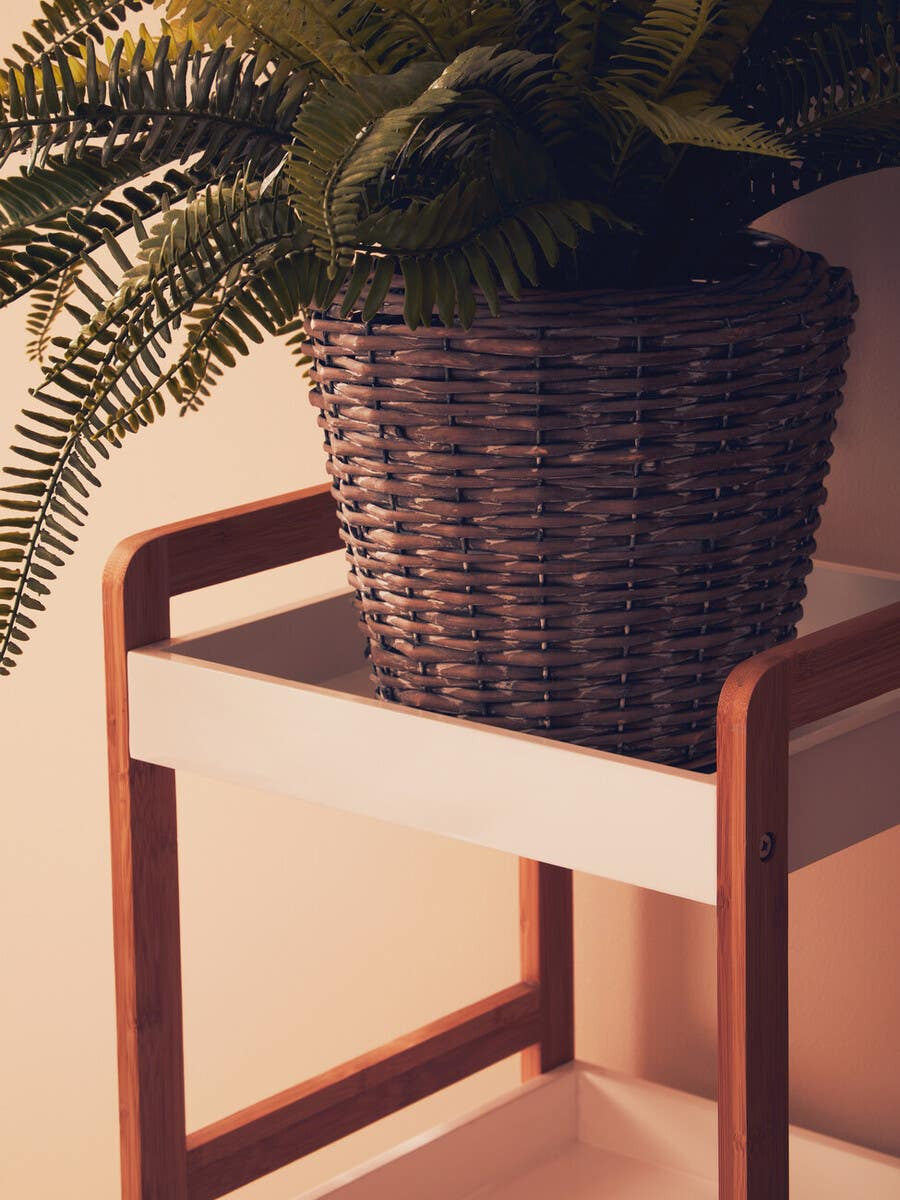 A close-up view of a four-tiered shelf unit featuring a white shelf with natural wood accents. A woven basket holds a lush green plant on the top shelf, showcasing a modern and minimalist design. The background is softly blurred, emphasizing the shelf unit's clean lines and natural materials