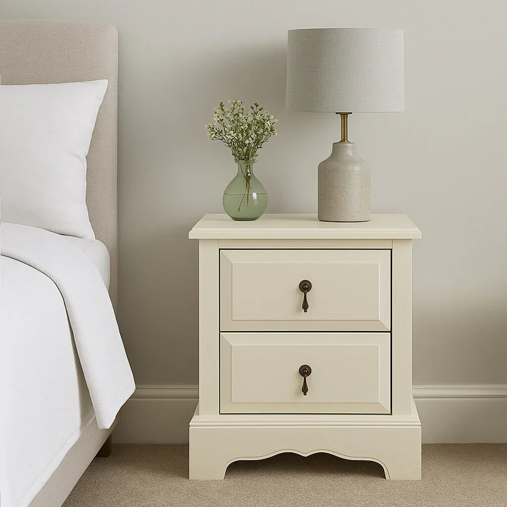 Cream bedside cabinet with two drawers, lamp, and vase beside bed in neutral bedroom