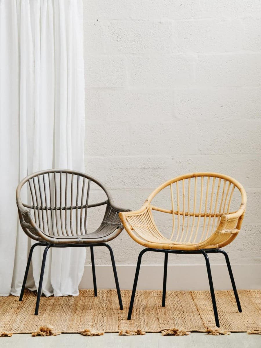 A grey wash natural rattan chair with a rounded backrest and open weave design, paired with a light-colored rattan chair, both resting on a wooden floor. A white curtain hangs in the background against a neutral wall