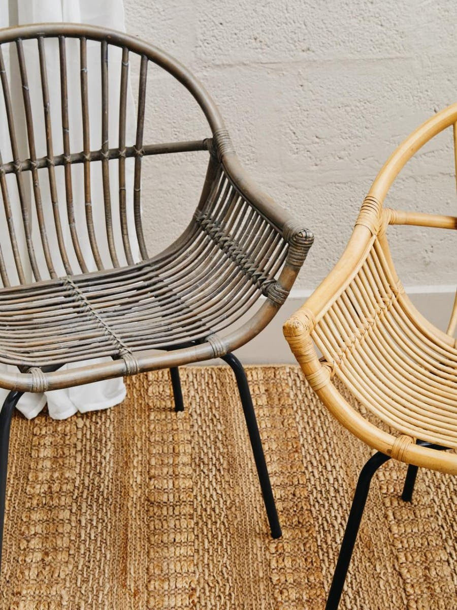 A grey wash natural rattan chair with a curved backrest and open design, positioned next to a lighter rattan chair, both resting on a textured woven rug. The background features a neutral wall, enhancing the natural tones of the chairs