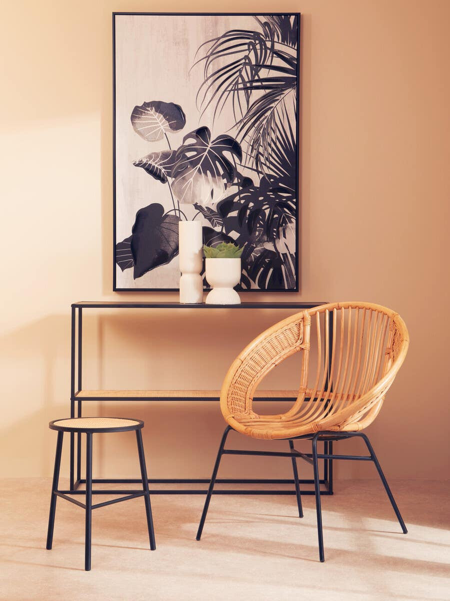 A natural rattan chair with a curved backrest and black iron legs is positioned next to a small black iron table. The scene features a large framed artwork with tropical leaves on a peach-colored wall, accompanied by two candles and a small plant on the table. A second stool is visible beside the chair