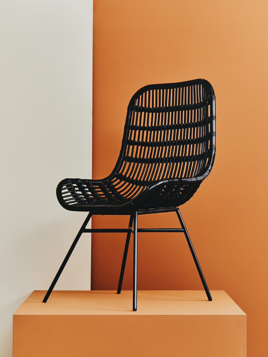 Curved black natural rattan chair with a modern design, featuring a slatted backrest and seat, supported by slender metal legs, displayed against a vibrant orange background
