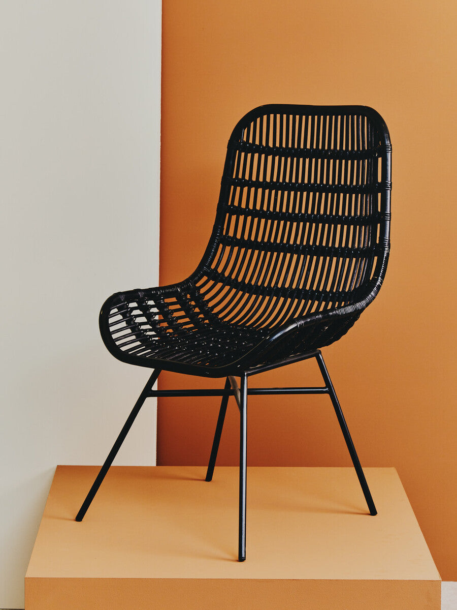 Curved black rattan chair with a natural finish, featuring a slatted design and slender metal legs, displayed against an orange backdrop. The chair has a modern aesthetic, emphasizing comfort and style