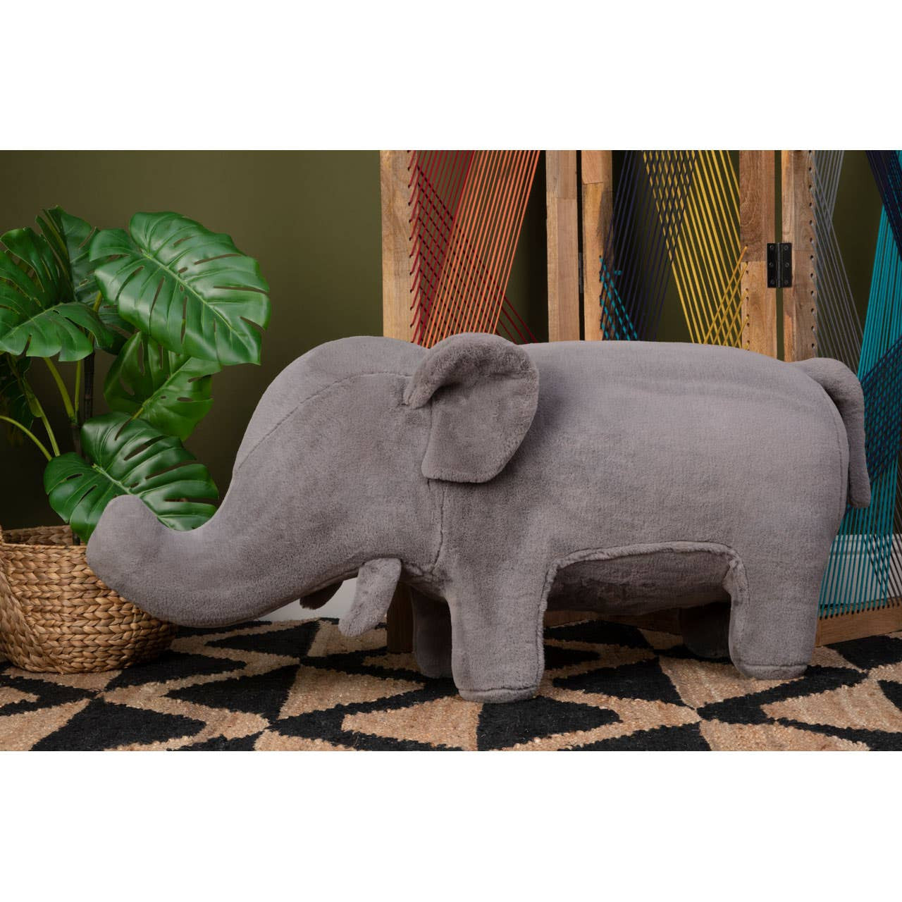 A grey elephant-shaped stool designed for kids, featuring a soft texture and rounded edges. The stool is positioned on a patterned rug with a plant in a woven basket nearby, set against a backdrop of wooden shelves