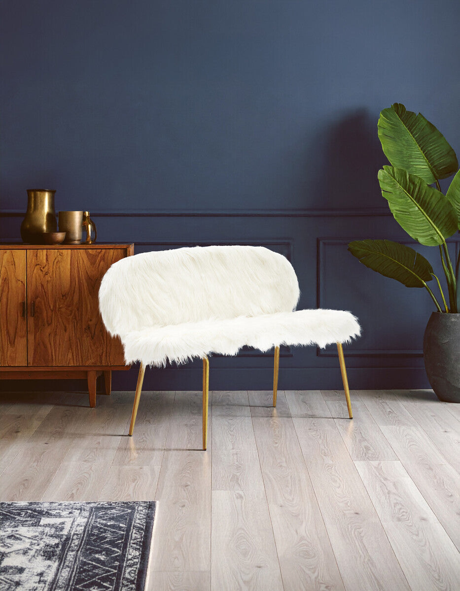Sienna white faux fur two-seat sofa with a curved backrest and gold finish legs, positioned against a dark blue wall. A wooden sideboard with decorative items is visible in the background, along with a large green plant and a patterned rug on the floor