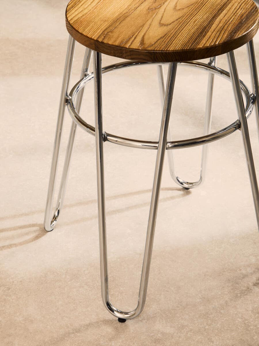 A wooden stool with a round top featuring a natural wood grain finish, supported by sleek metal hairpin legs. The legs are thin and tapered, providing a modern and minimalist look. The stool is positioned on a light-colored floor, highlighting its design