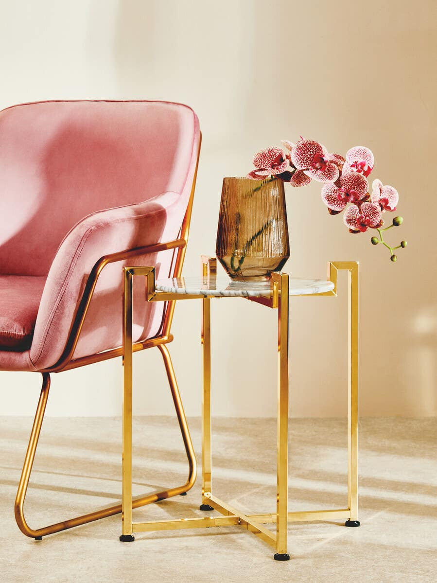 A white marble side table with a warm gold base, positioned next to a pink upholstered chair. The table features a sleek, modern design and holds a decorative vase with pink flowers. The background is softly lit, enhancing the elegant aesthetic of the scene