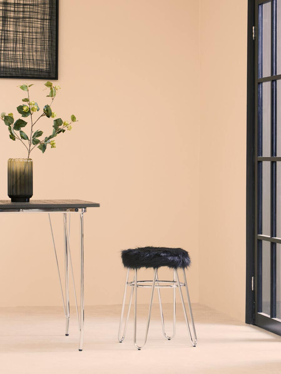 A black faux fur stool with a sleek design, positioned next to a modern glass table. The background features a light beige wall and a large window with black frames, while a vase with flowers sits on the table, adding a touch of greenery to the scene