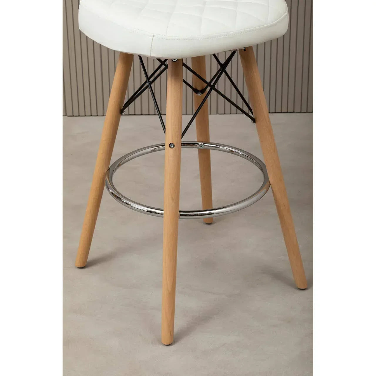 Bar stool with light wooden legs, chrome footrest, and white padded seat on beige floor