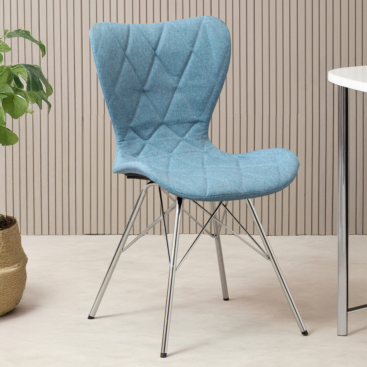 A stylish blue dining chair with a quilted fabric seat and backrest, supported by sleek metal legs, positioned next to a small round table and a potted plant in a modern interior setting