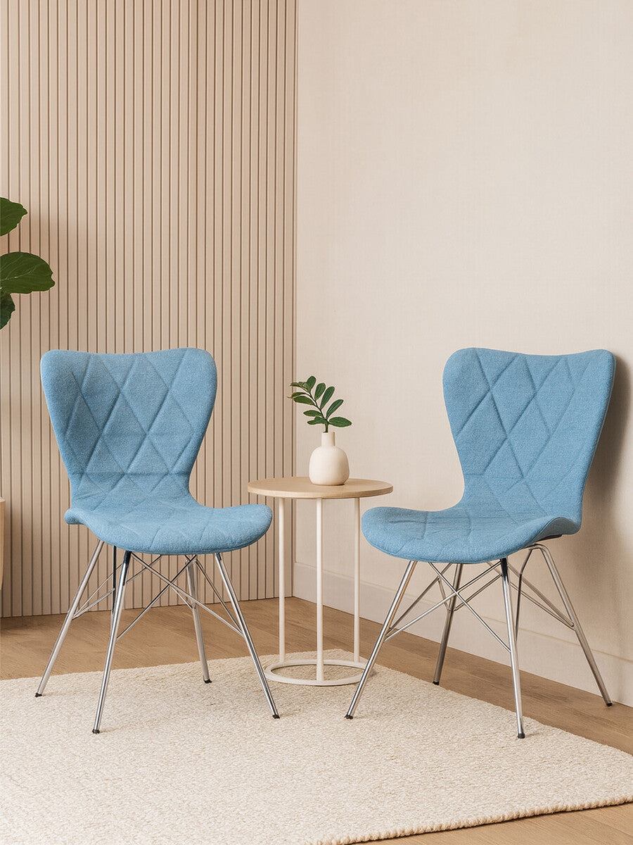 Two Stockholm blue dining chairs with a quilted design are positioned around a small round table. A potted plant sits on the table, and the background features light-colored walls with subtle texture. A soft area rug is visible beneath the chairs