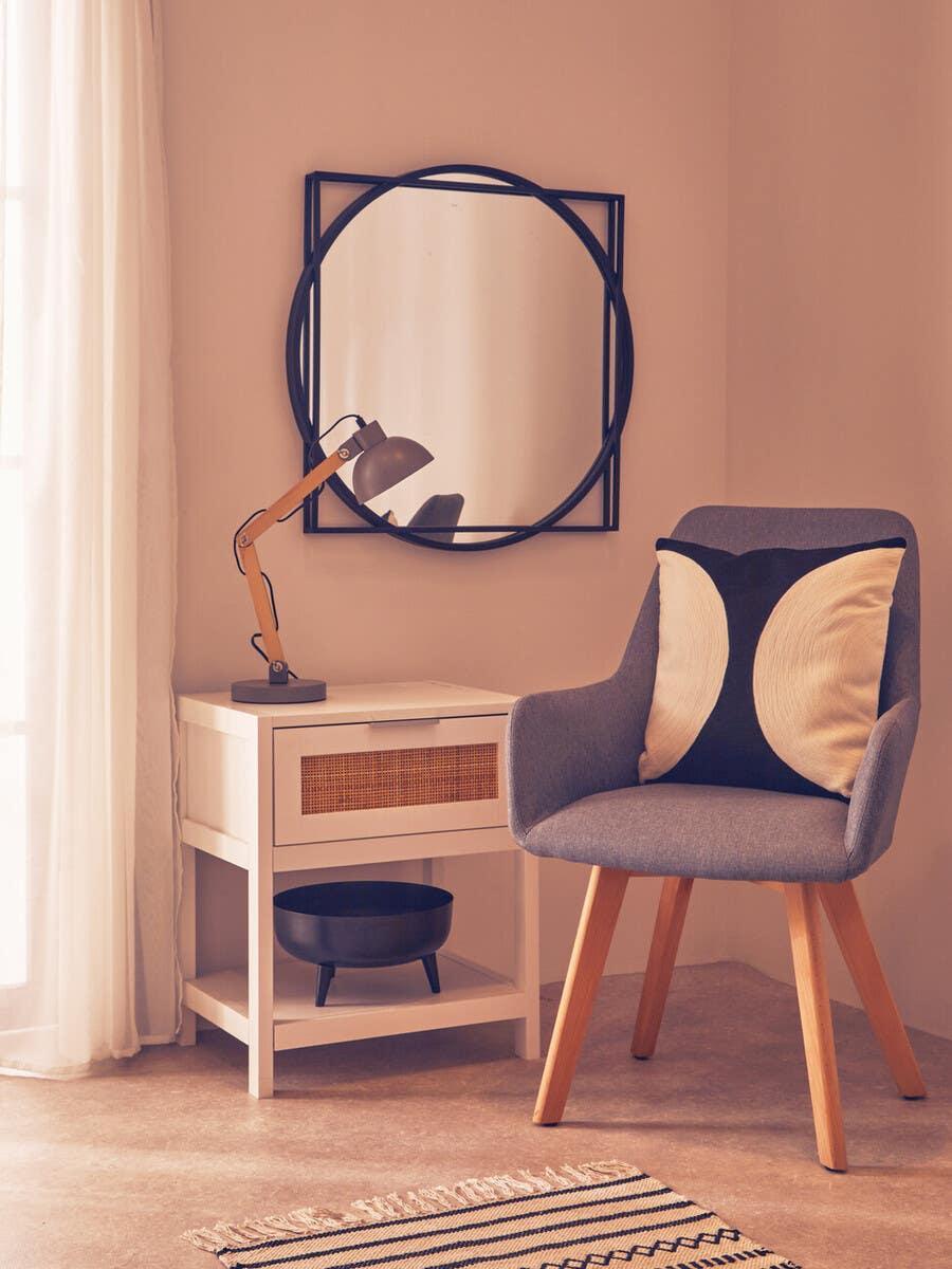 Grey armchair with cushion, white bedside table, black lamp and round framed wall mirror