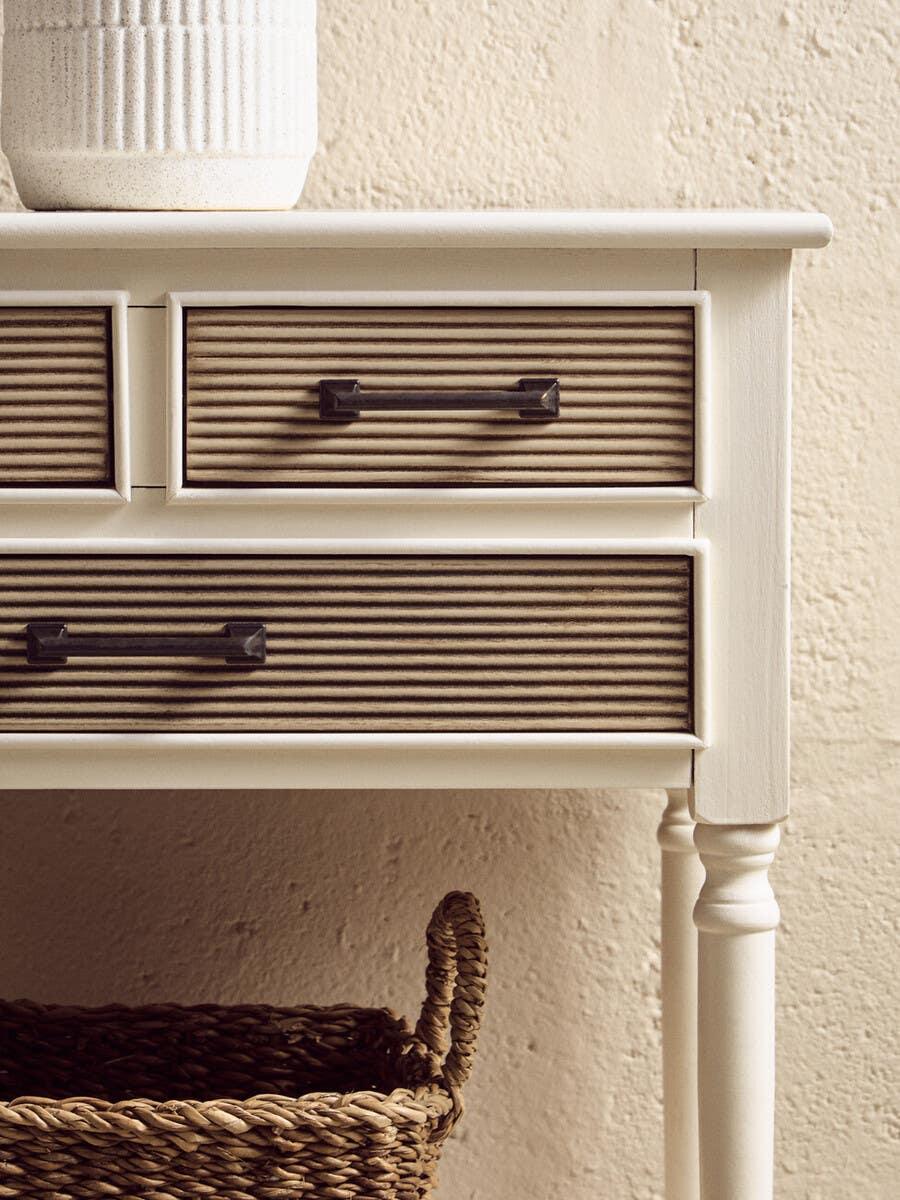 White wooden console table with grooved drawers and black handles, wicker basket below