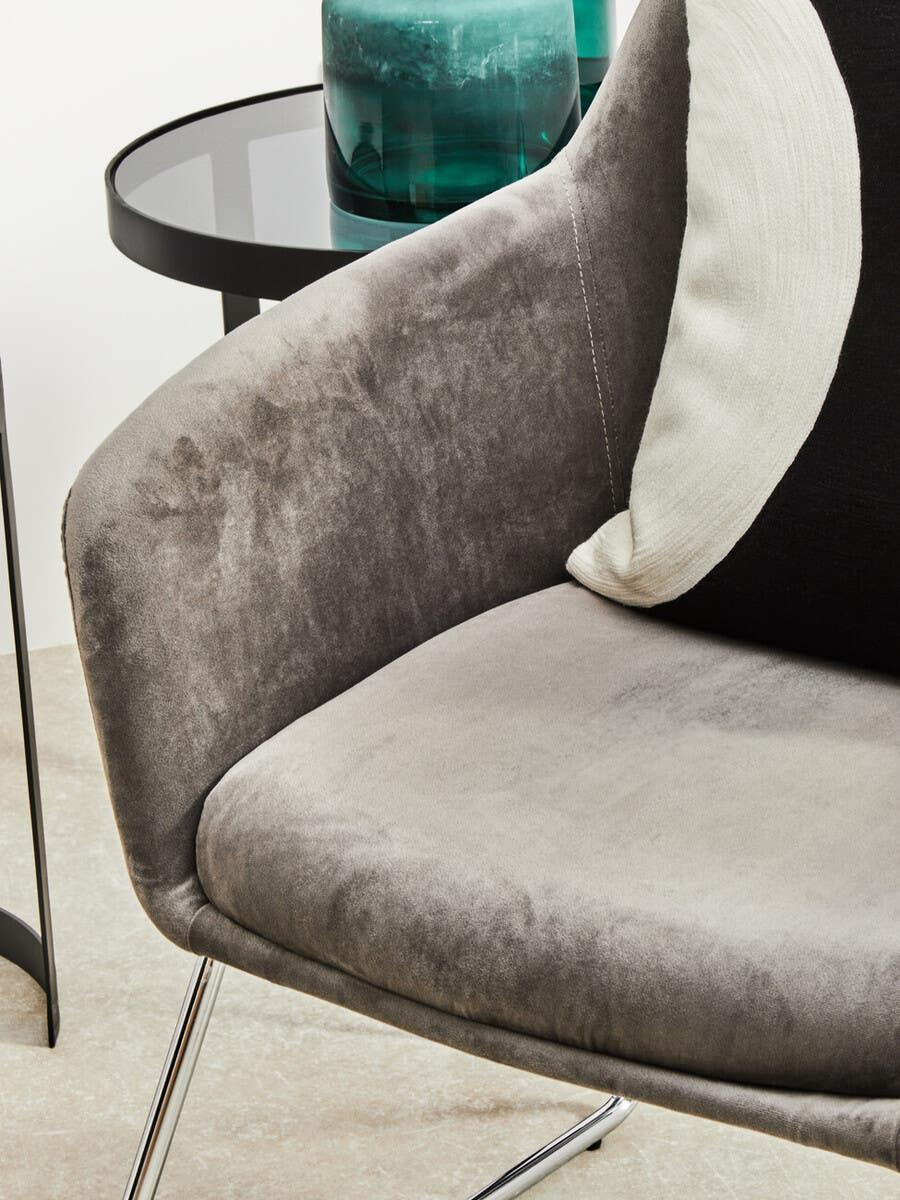 Grey velvet armchair with chrome legs beside black glass side table and green glass vase