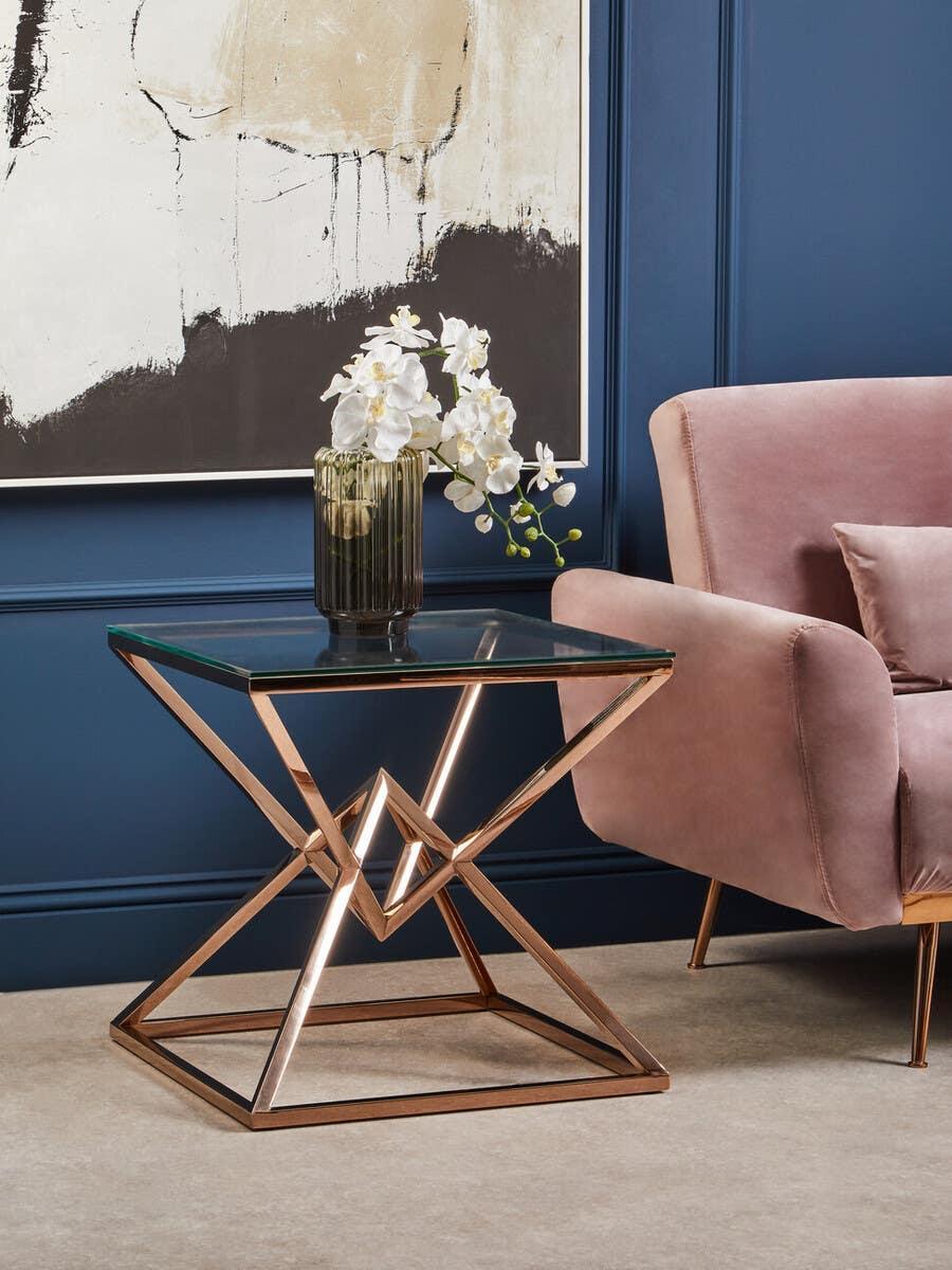 Glass top side table with gold metal geometric frame beside pink velvet armchair