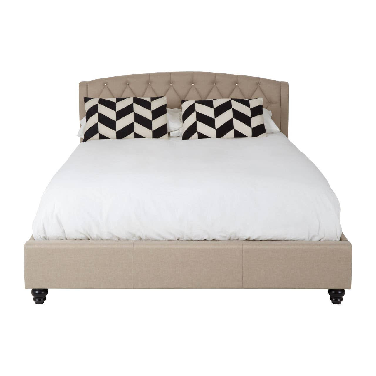 Beige king bed with a tufted headboard, featuring a white mattress and decorative black and white chevron-patterned pillows. The bed is supported by dark wooden legs