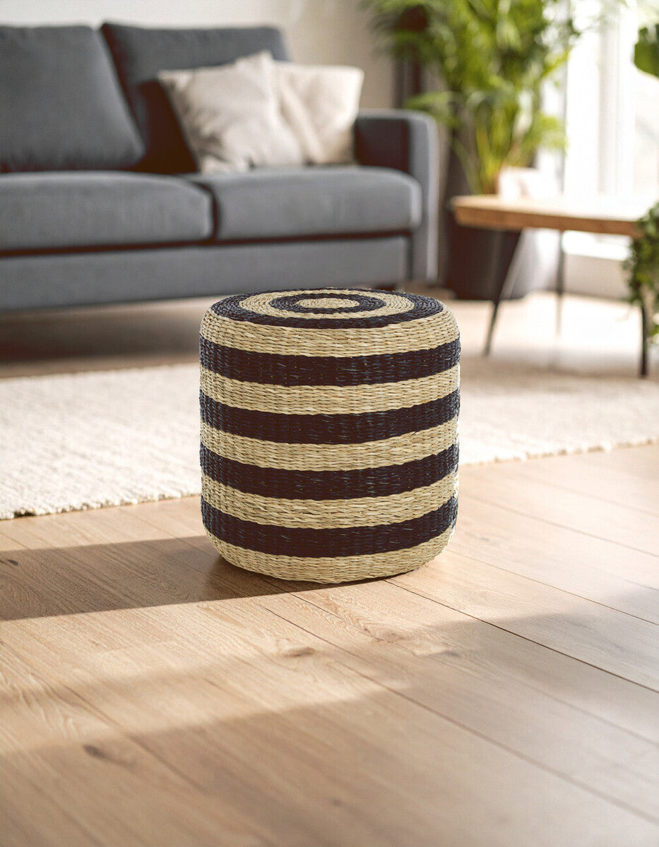 Round pouffe made of seagrass featuring alternating black and natural stripes, placed on a light wooden floor in a bright room with a sofa and decorative pillows in the background