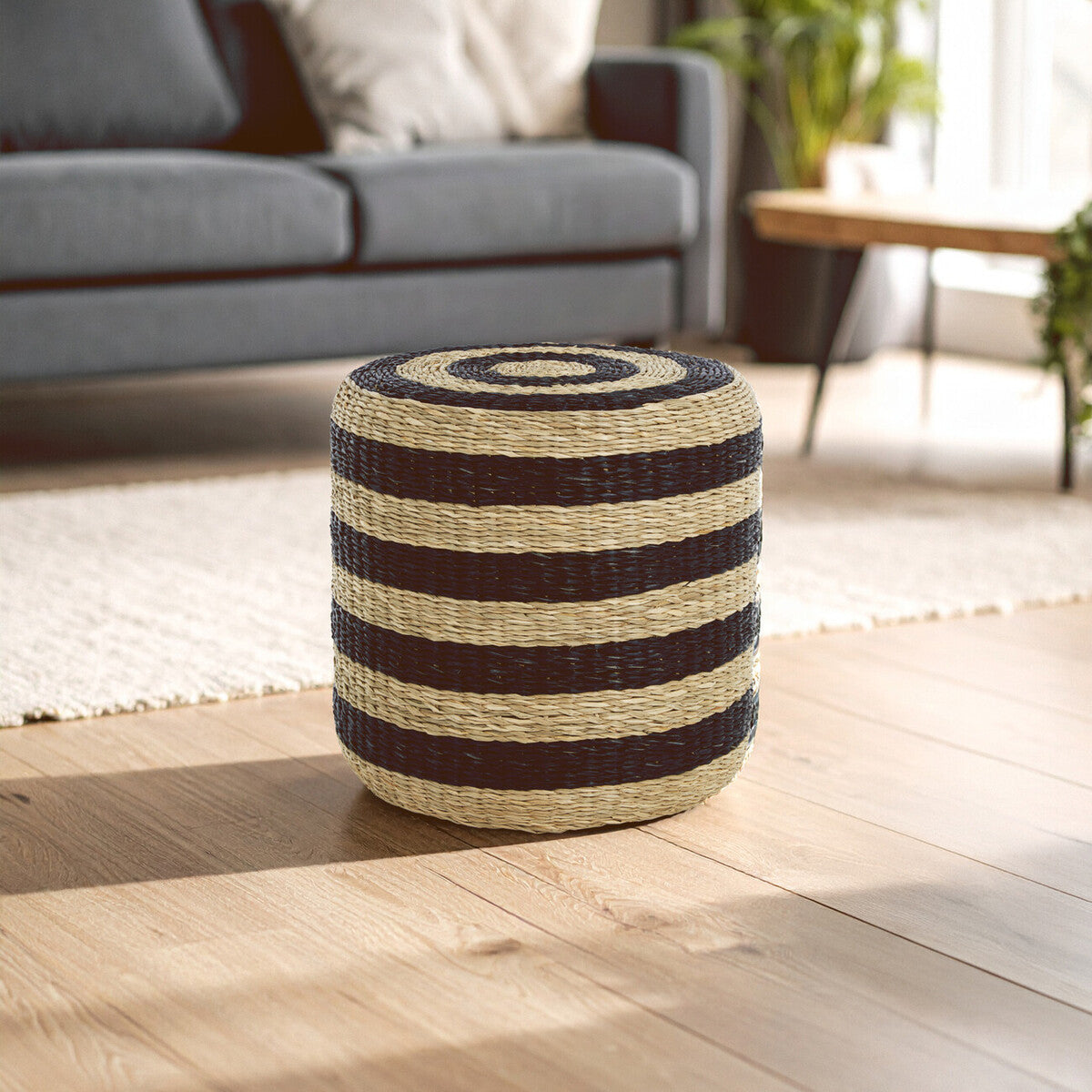 Round pouffe made of seagrass featuring alternating black and natural-colored stripes, placed on a light wooden floor in a bright room with a gray sofa and a large window in the background