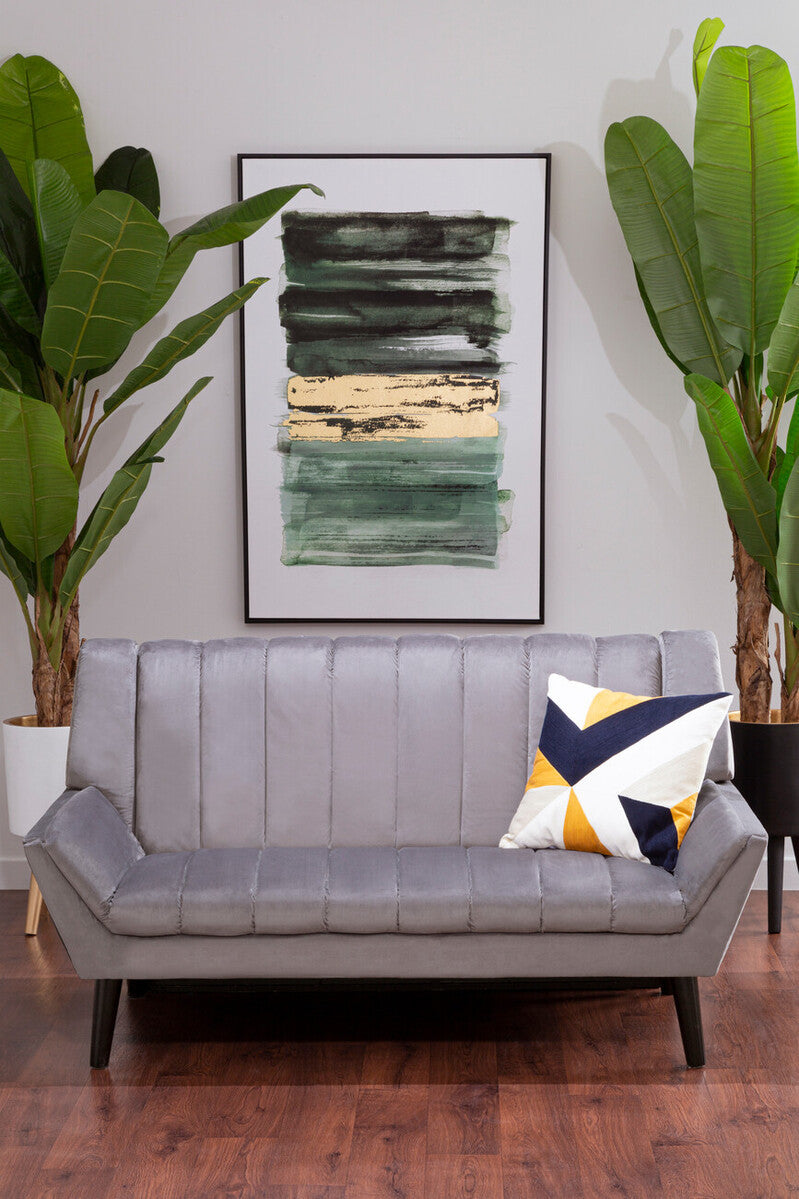 A grey two-seat sofa with a tufted design, positioned in a modern living space. The sofa features black legs and is adorned with a colorful geometric throw pillow. Behind the sofa, there is a large framed abstract artwork and two tall green plants, creating a stylish and inviting atmosphere. The floor is made of warm wood, adding to the cozy ambiance