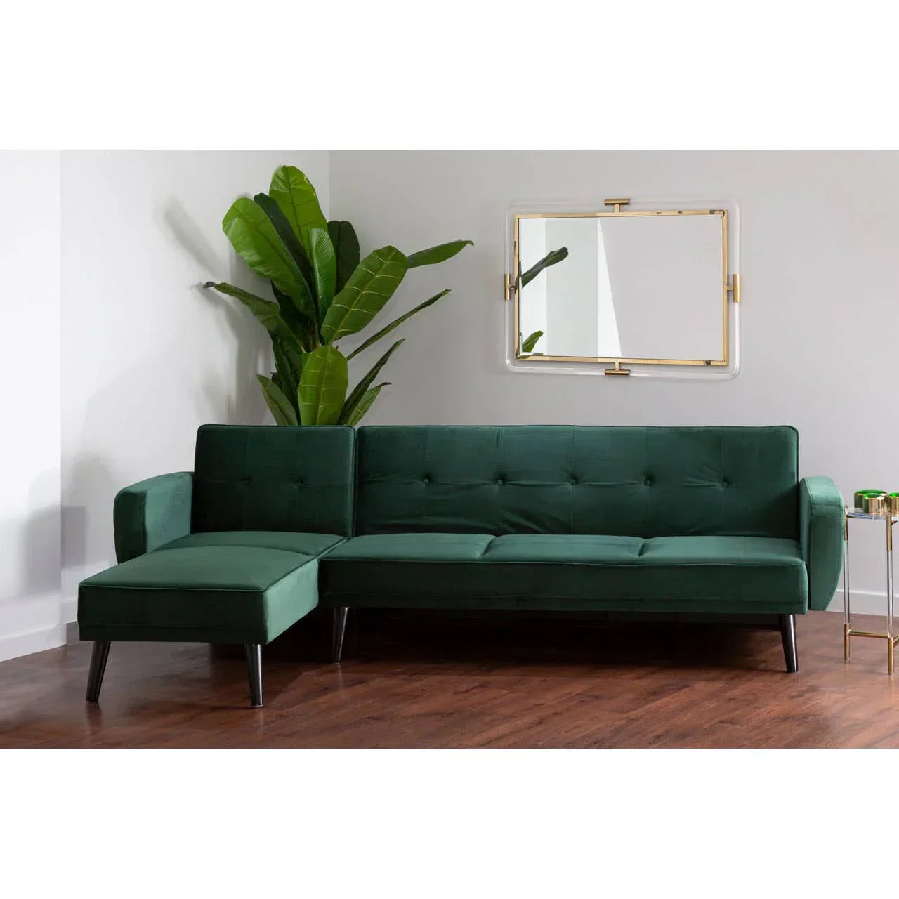 Green velvet corner sofa with chaise on wooden floor near plant and wall mirror