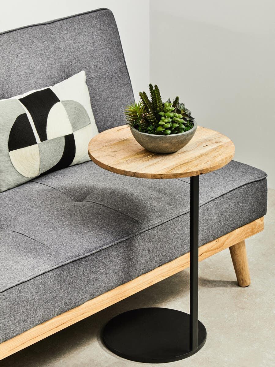 Round wooden side table with black metal base beside grey fabric sofa and cushion