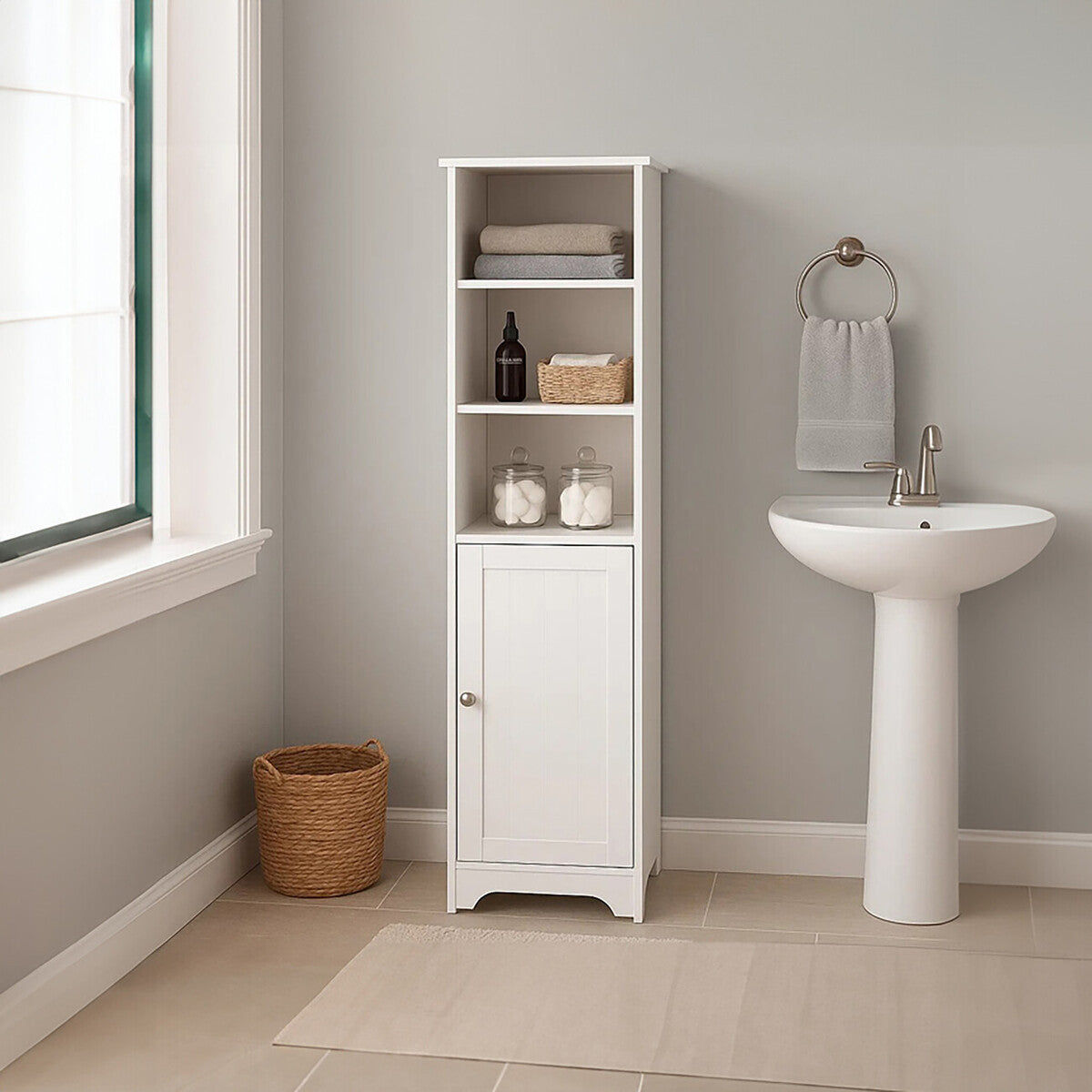 A tall, white single door cabinet with two open shelves and a closed lower compartment, positioned next to a white pedestal sink in a light-colored bathroom. A woven basket is placed on the floor beside the cabinet, and natural light streams in through a window