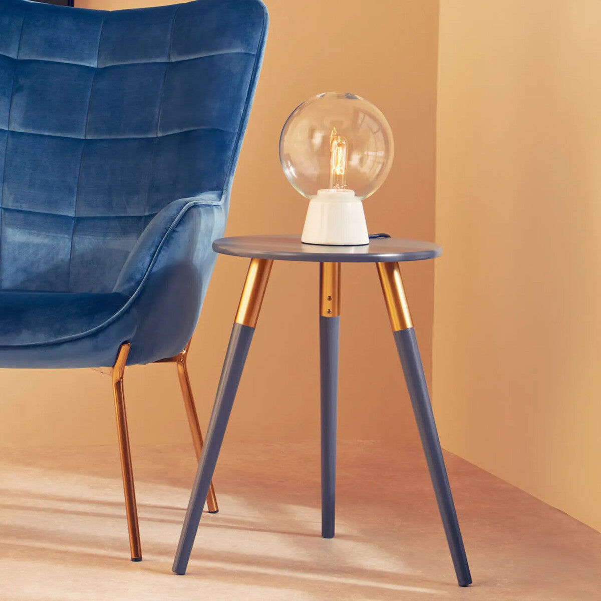 A dark grey side table with a smooth surface and three tapered legs, two of which are gold-colored. A stylish lamp with a round glass shade sits on top of the table, next to a blue upholstered chair against a warm-toned wall