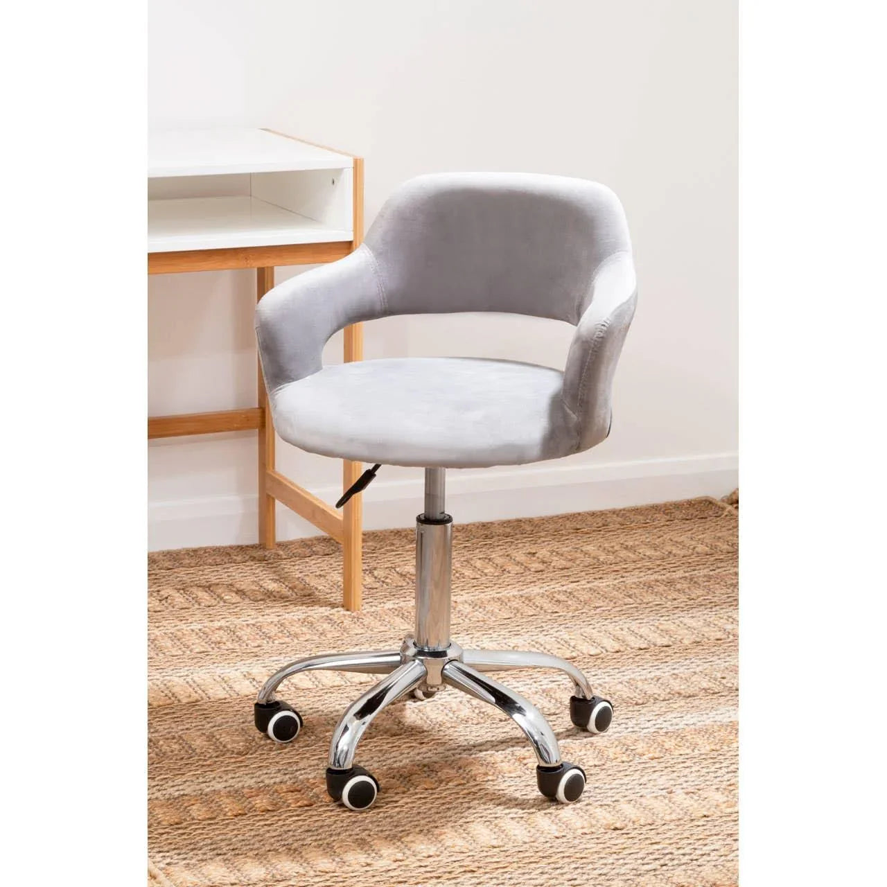 Grey swivel office chair with chrome base and wheels on woven rug beside wooden desk
