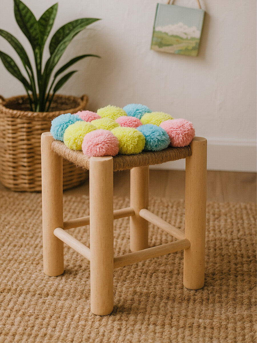 A wooden stool with a natural finish features a colorful top adorned with pink, blue, and yellow pom poms. The stool has four sturdy legs and is placed on a textured beige rug, with a woven basket and a small piece of artwork visible in the background